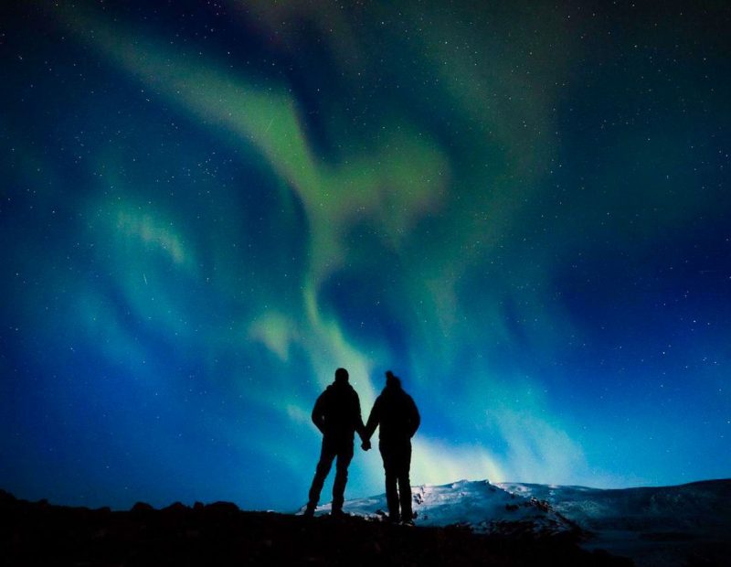 a couple holding hands and standing on a hilltop, looking at the Northern Lights