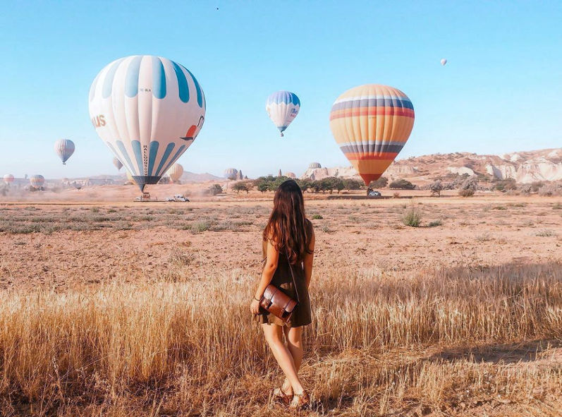 a girl watching a group of hot air balloons flying over a field of tall grass